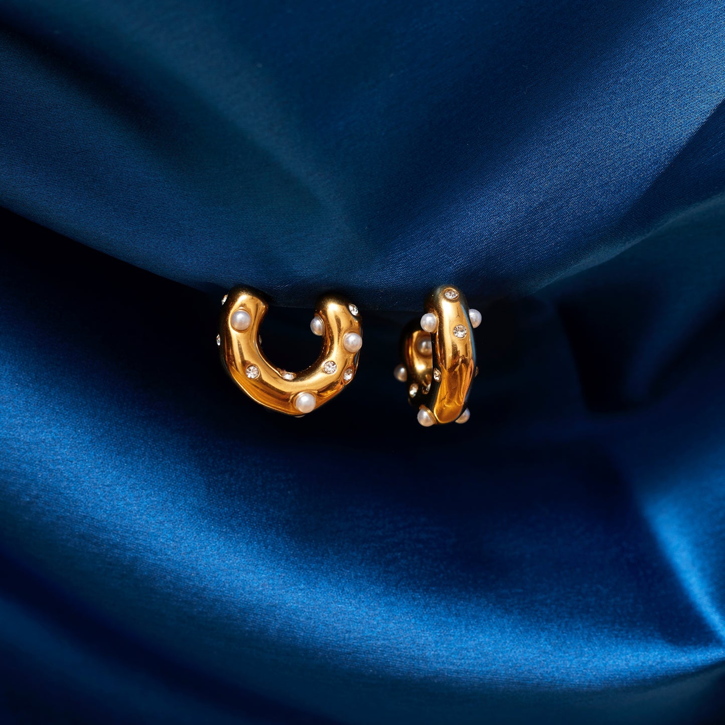 A close-up image of a pair of gold-plated hoop earrings with pearls and small sparkling stones, displayed against a blue fabric backdrop.