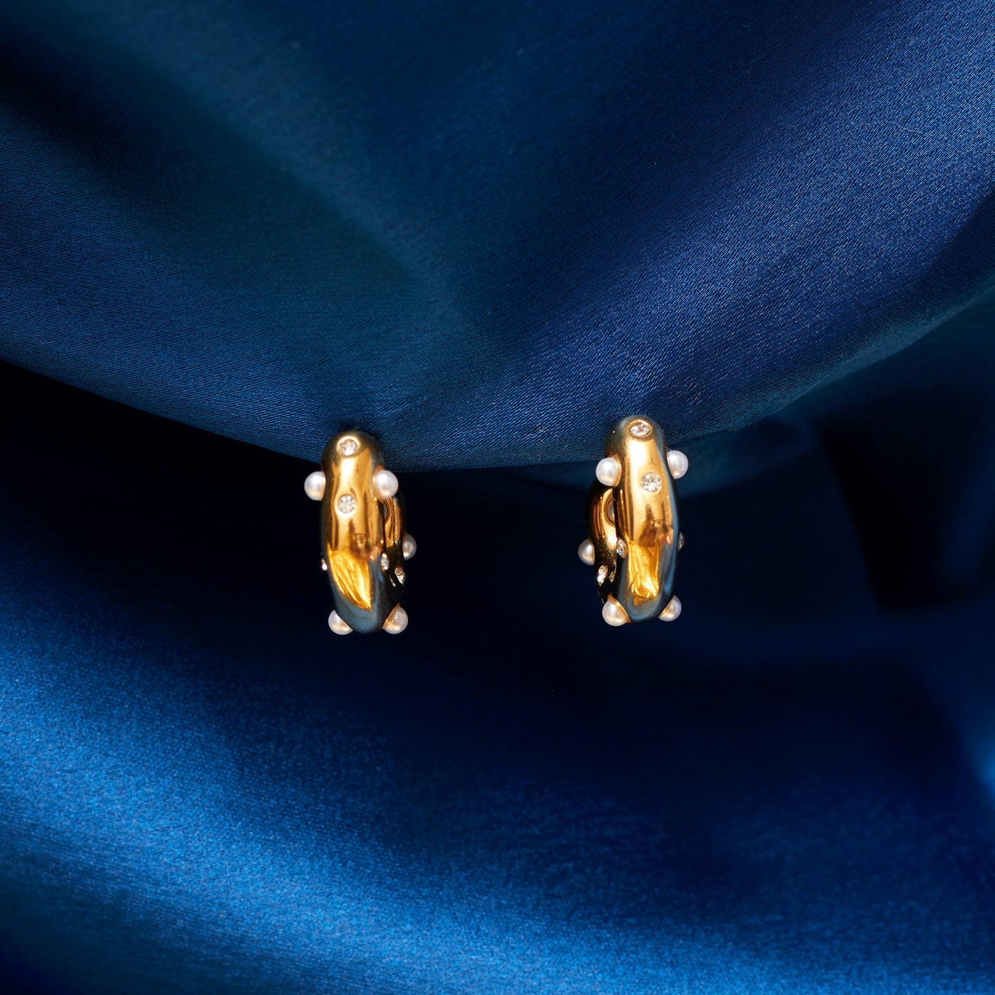 A close-up image of a pair of gold-plated hoop earrings with pearls and small sparkling stones, displayed against a blue fabric backdrop.