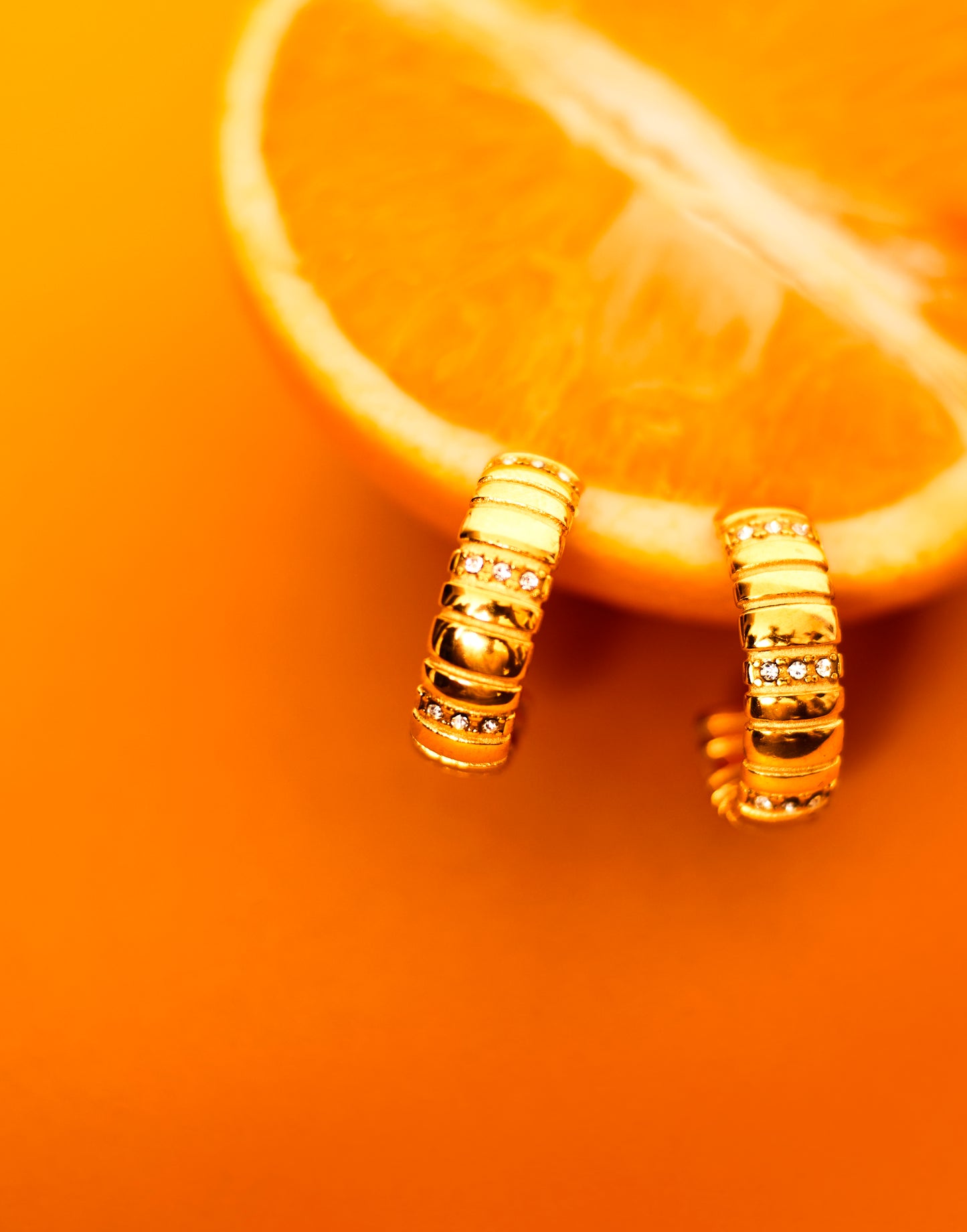 A close-up image of a gold-plated chunky hoop earring placed on a sliced orange, with a plain orange background.