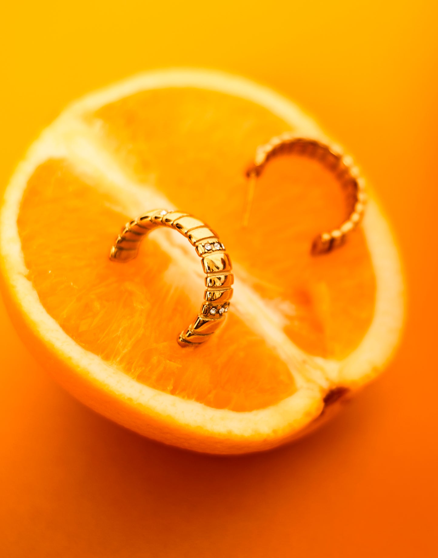 A close-up image of a gold-plated chunky hoop earring placed on a sliced orange, with a plain orange background.