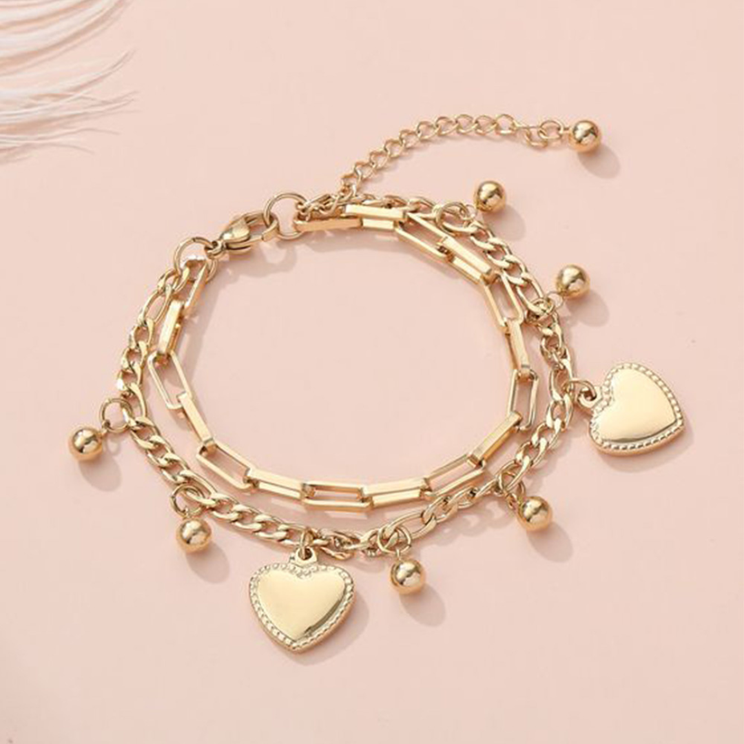 A gold-colored layered bracelet featuring heart charms and small bell-like embellishments on a pink background.