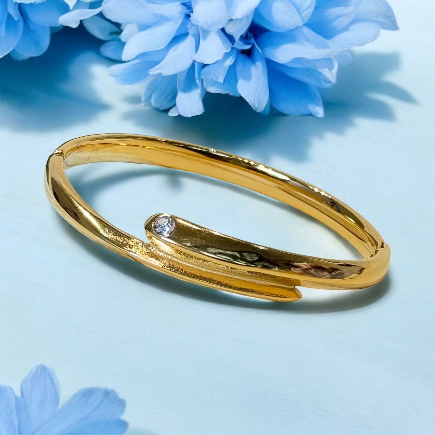 A gold-plated, curved bracelet with a central stone, displayed on a background with blue flowers.
