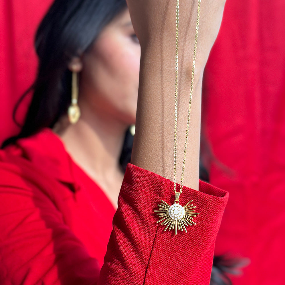 A person wearing a gold-plated necklace with a half sun enamel pendant, held up against a red background.