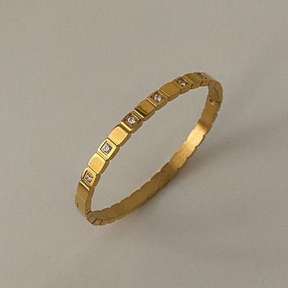A gold-plated bracelet with a unique design that is bold at the center and tapers towards the clasp, displayed against a grey background.