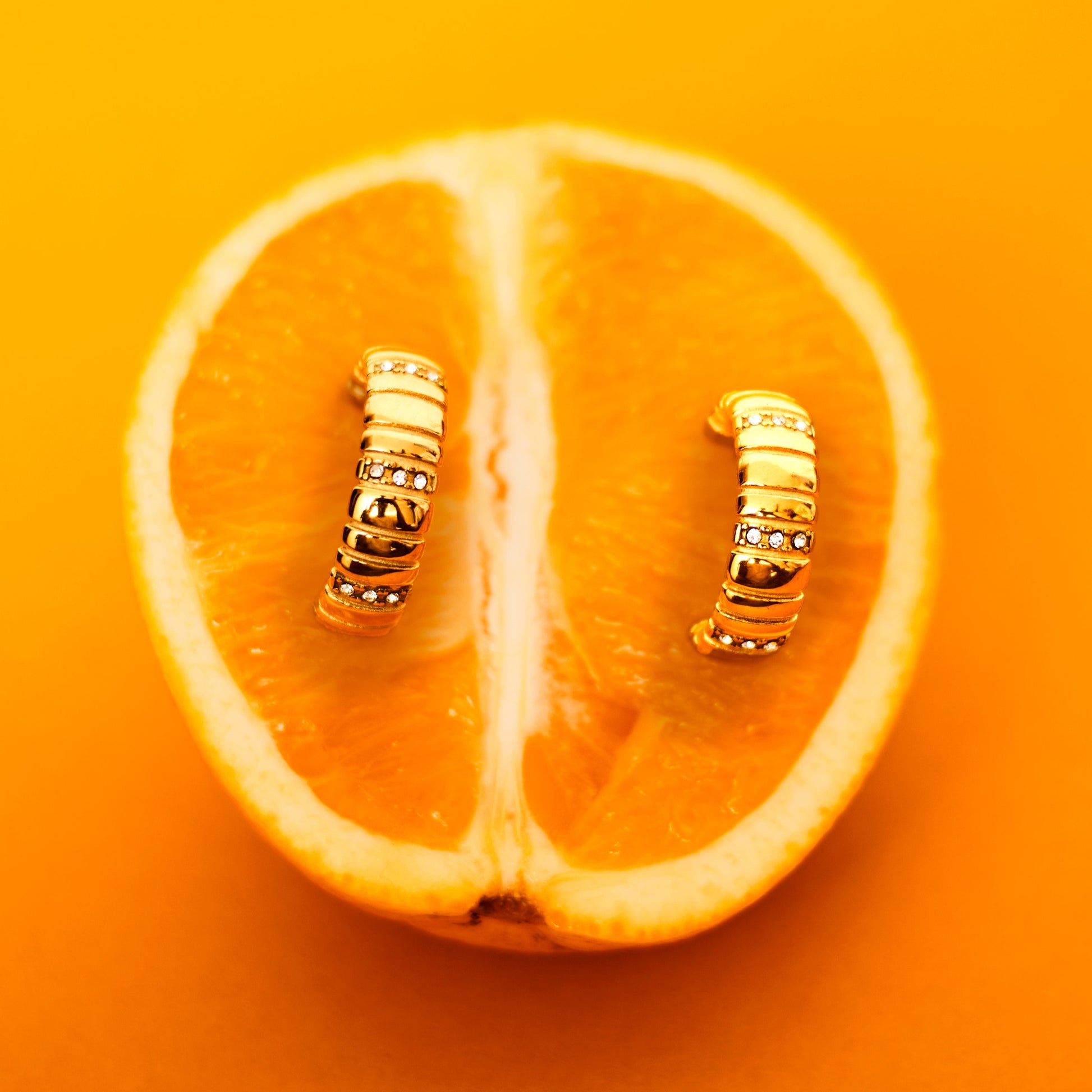 A close-up image of a gold-plated chunky hoop earring placed on a sliced orange, with a plain orange background.