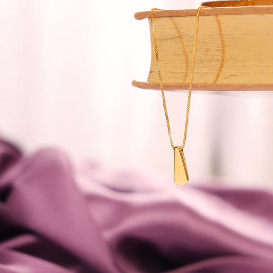 A gold-plated drop pendant necklace hanging over a book, with a blurred purple fabric in the background.