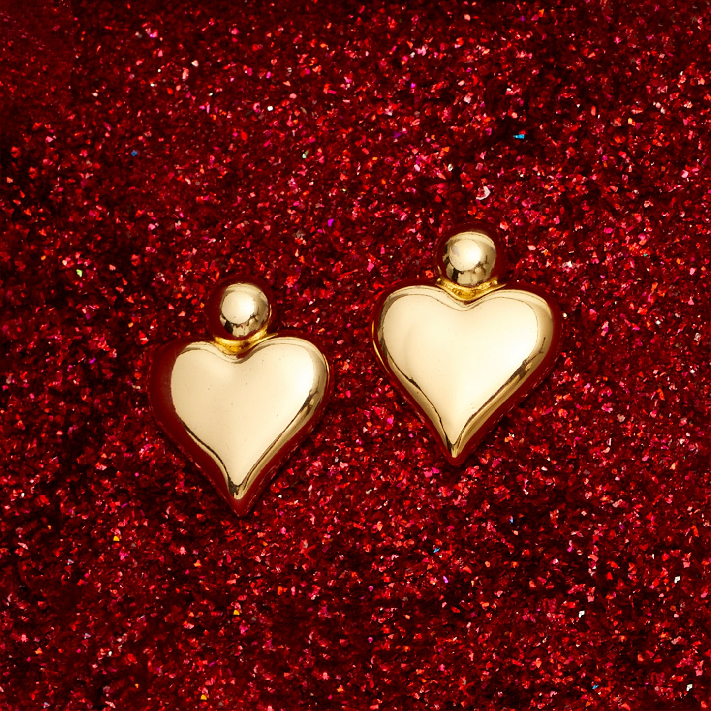 A pair of 18K gold plated heart-shaped earrings displayed against a red glittery background.