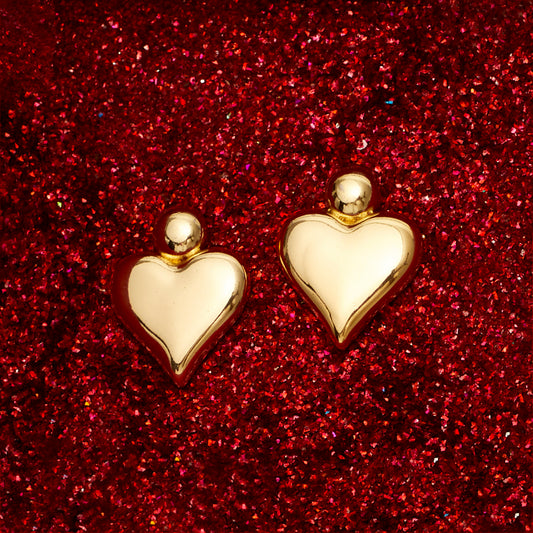 A pair of 18K gold plated heart-shaped earrings displayed against a red glittery background.