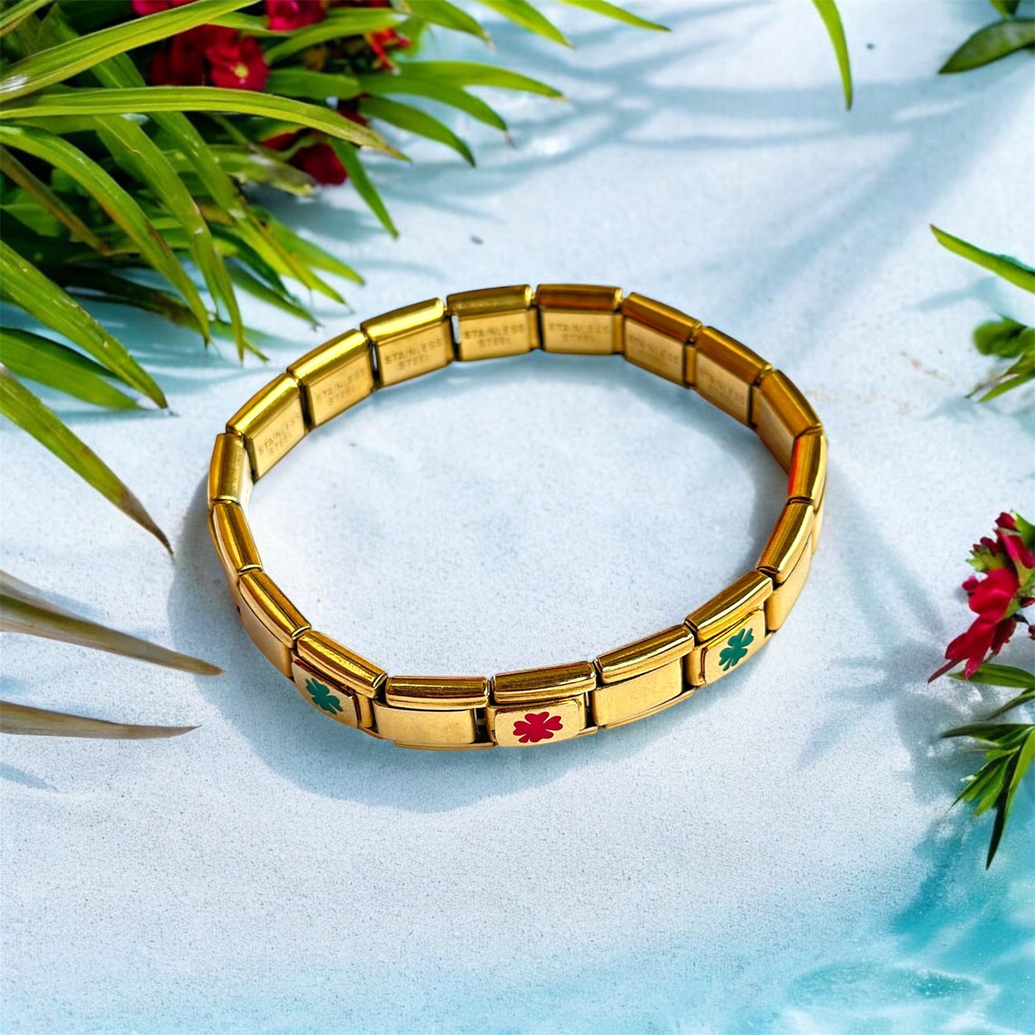A gold-colored Italian charm bracelet with colorful charms, displayed on a blue surface with green leaves and red flowers in the background.