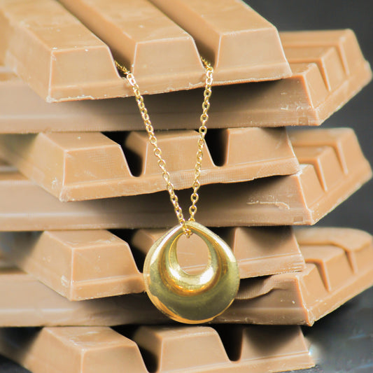A gold-toned necklace with petal shaped pendant , displayed against a chocolate bar background.